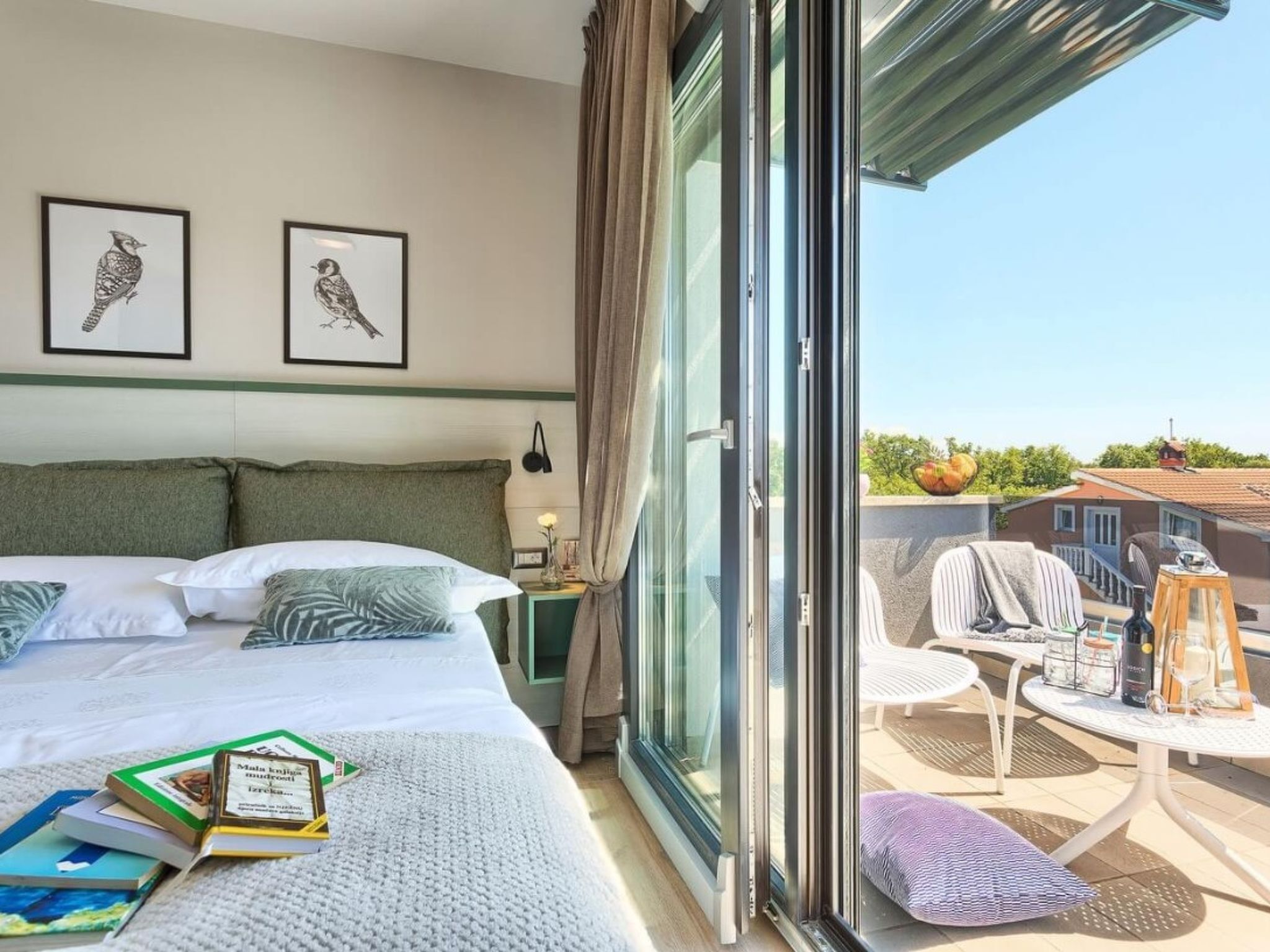 A hotel room with a bed, a side table, a chair, a balcony with a table, and two framed pictures on the wall.