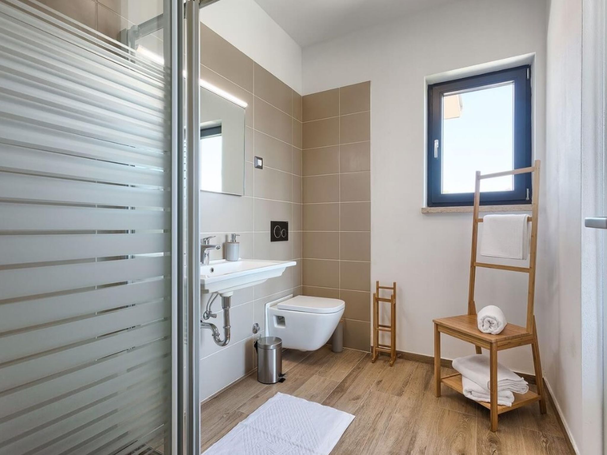 A modern bathroom with a glass shower enclosure, a sink, a toilet, a wooden ladder with towels, and a window.