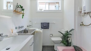 A compact bathroom with a bathtub, toilet, and sink. There is a window above the toilet, a towel rack, and a plant. - Popular North Cronulla Beach Apartment sleeps up to 5 people (Cronulla)