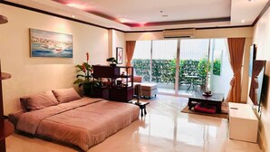 A modern hotel room with a large bed, a small desk, a chair, a TV, and a balcony with a view of greenery. - Japanese Styled Condo in the Heart of Pattaya next to Pattaya Beach. (Pattaya City)