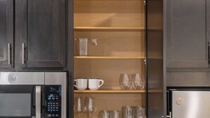 Een keuken met donkere kastjes, een magnetron, een koelkast en een plank met bekers, glazen en een maatbeker.