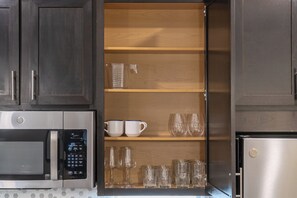 Eine Küche mit dunklen Schränken, einer Mikrowelle, einem Kühlschrank und einem Regal mit Tassen, Gläsern und einem Messbecher.