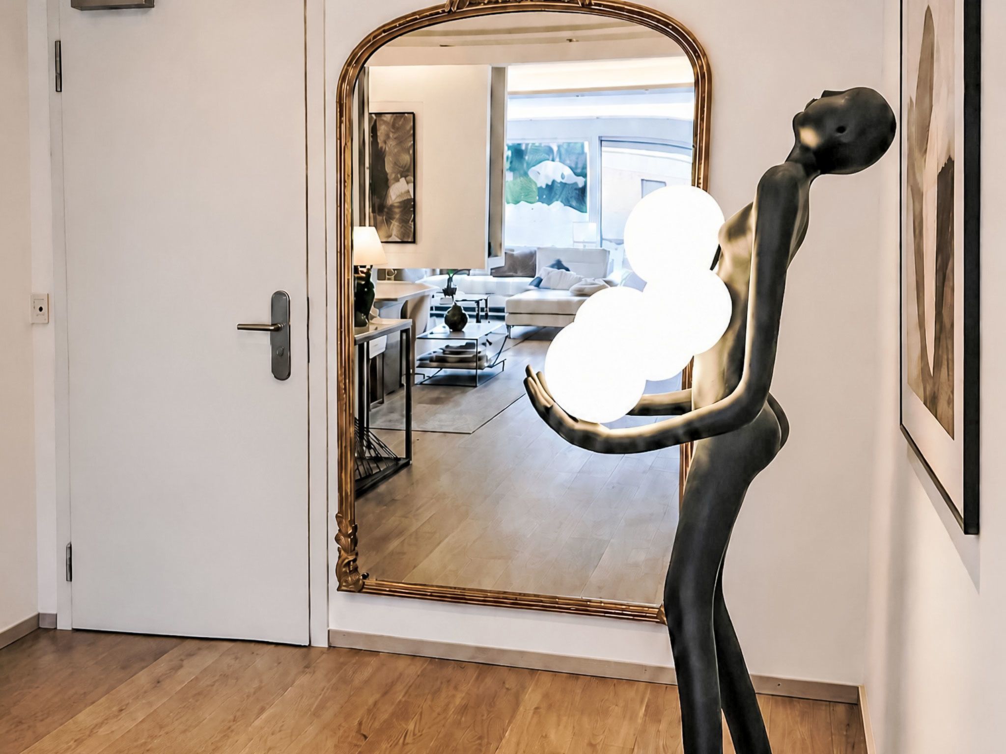 Ein Zimmer mit einem großen Spiegel, der einen Wohnbereich reflektiert, einer Tür mit moderner Griffleiste und einem hellen Holzboden.