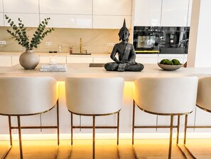 A modern kitchen with a central island, bar stools, a Buddha statue, and a potted plant.