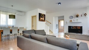 Ein modernes Wohnzimmer mit grauem Sofa, Holzboden, Essbereich mit Stühlen, einer Treppe, einem Fernseher und einem Gemälde an der Wand.