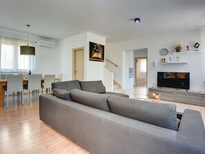 Ein modernes Wohnzimmer mit grauem Sofa, Holzboden, Essbereich mit Stühlen, einer Treppe, einem Fernseher und einem Gemälde an der Wand.