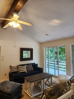 A living room with a ceiling fan, a dark-colored sofa, a wooden coffee table, and a large window with a view of greenery. - The Blue Dove (Shenandoah Farms)