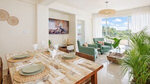 A modern living room with a dining area, featuring a large wooden table set for dining, green armchairs, a large window with a view of palm trees, and a hanging light fixture.