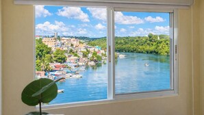 A room with a large window overlooking a river with boats and a densely built-up area with greenery and buildings.