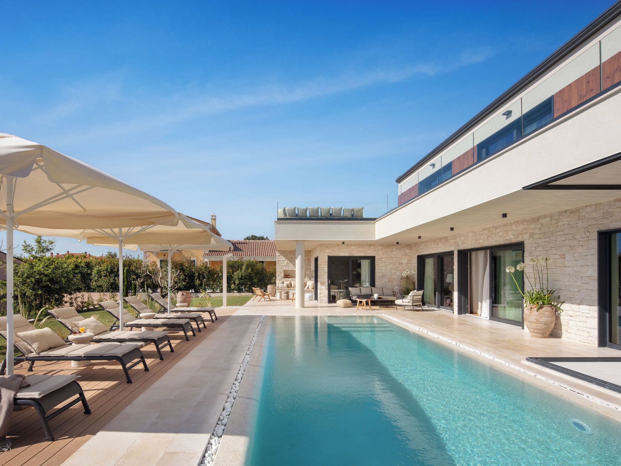 Uma área de piscina moderna com espreguiçadeiras e guarda-sóis, piscina azul límpida e um edifício de pedra com grandes janelas.