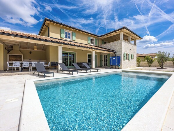 Un espace aménagé au bord de la piscine, avec des chaises longues, un barbecue et un bâtiment en pierre surmonté d’un toit en tuiles.
