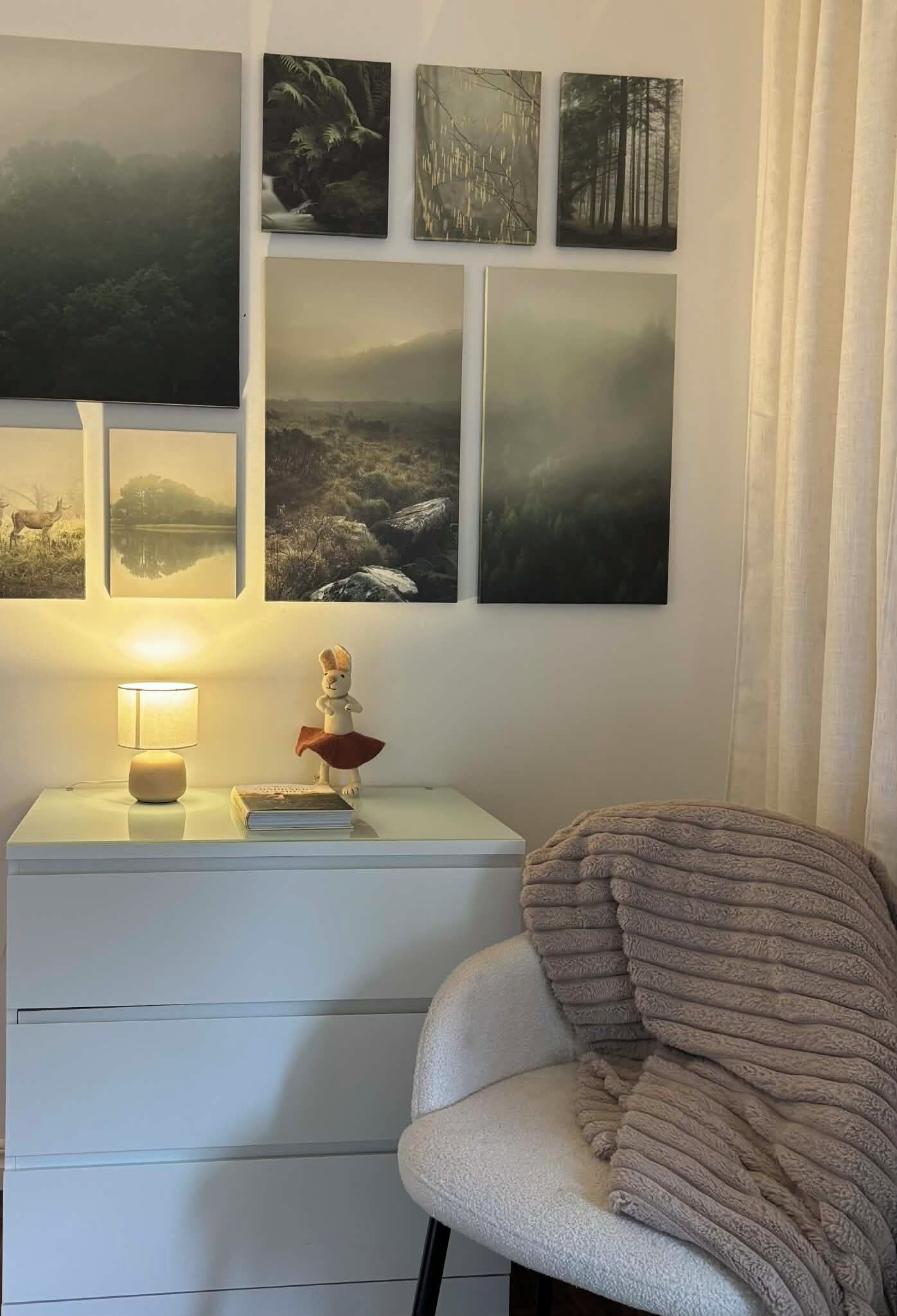 A bedroom with a white nightstand, a lamp, a plush chair, and a wall adorned with nature-themed artwork.