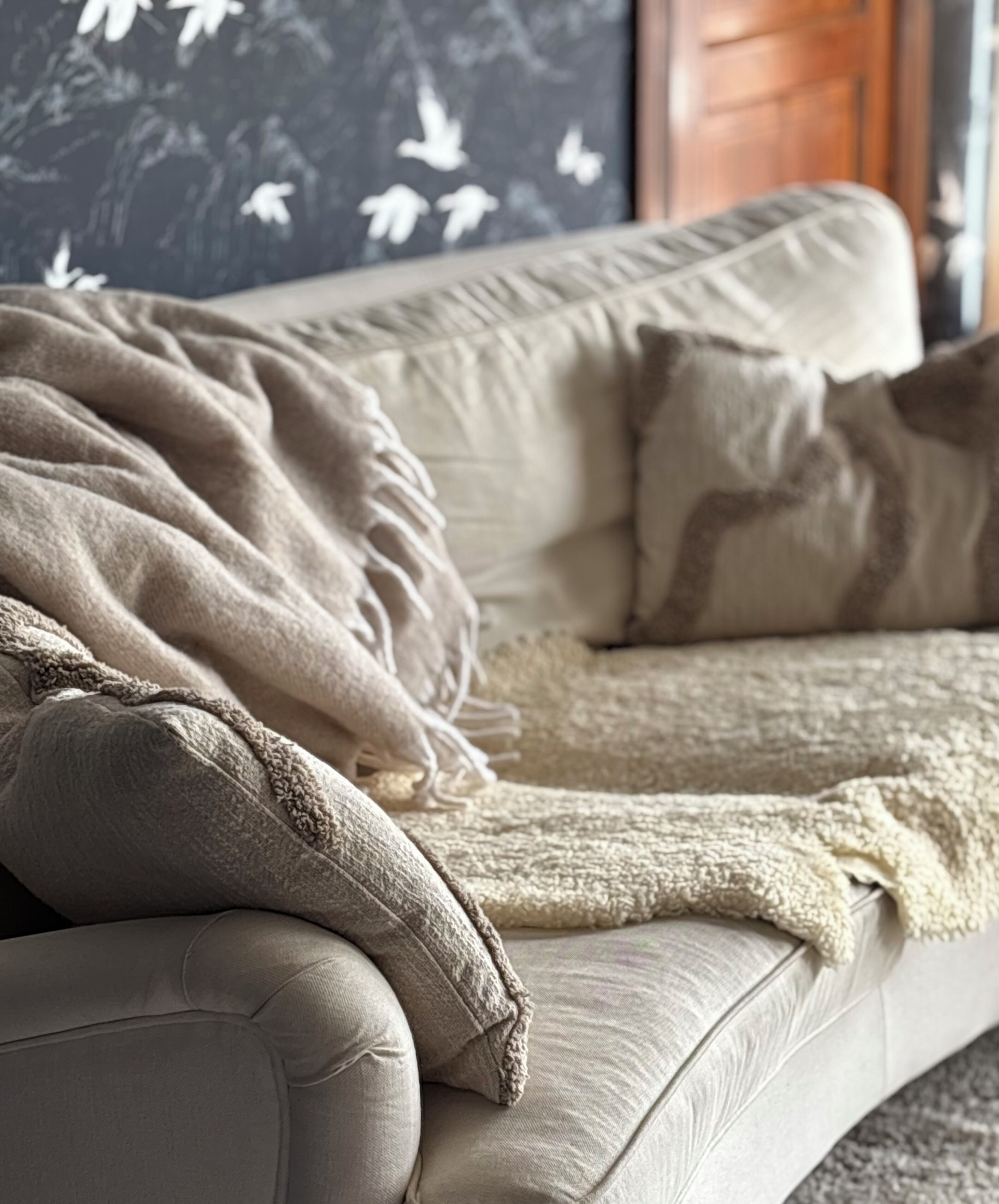 A beige sofa with a throw blanket and pillows, set against a wall with a floral pattern.