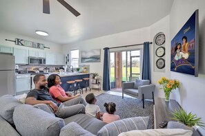 Ein modernes Wohnzimmer mit einer grauen Couch, einem Couchtisch und einem Fernseher. Im Hintergrund ist eine Küche mit weißen Schränken und einer Mikrowelle zu sehen.