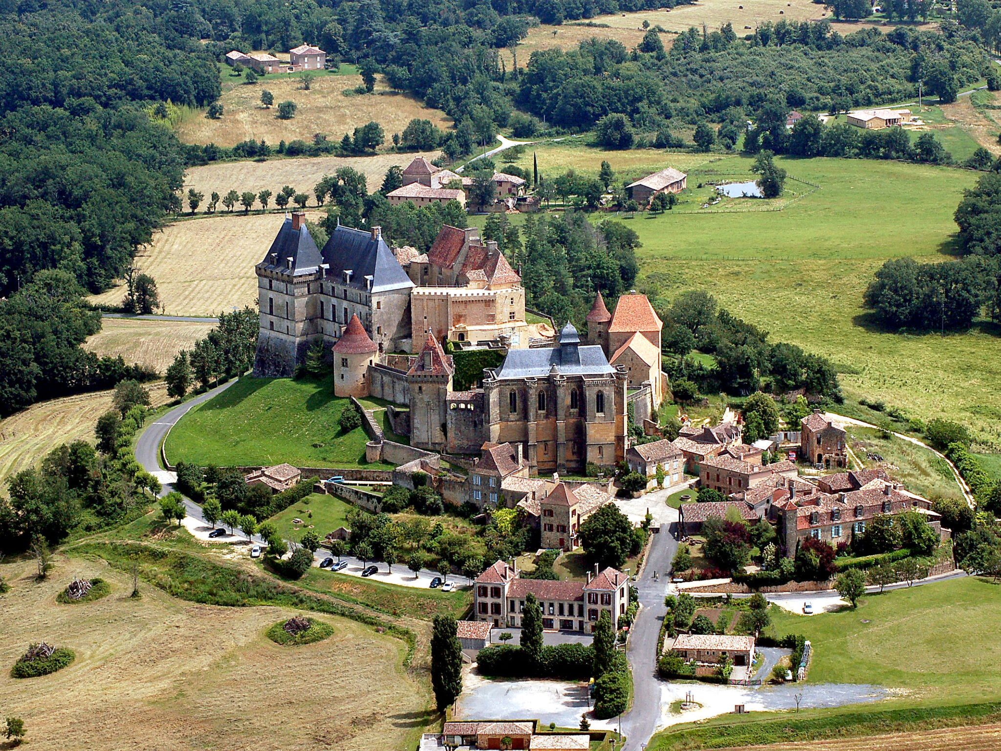 Une vue aérienne d’un ensemble castral historique comprenant plusieurs bâtiments, dont une grande structure centrale surmontée d’une tour, le tout étant entouré de maisons plus petites et de champs.