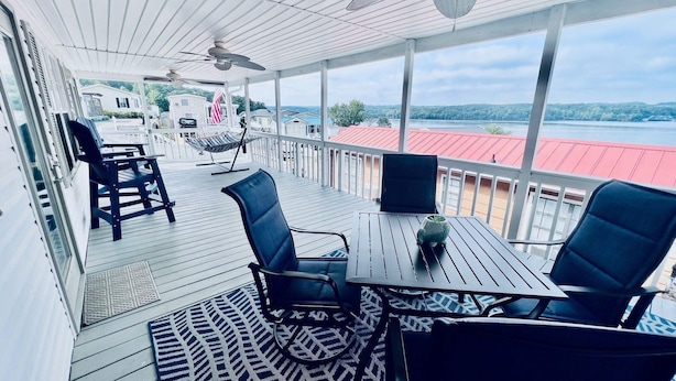 Une terrasse extérieure couverte, dotée d’une table et de chaises, d’un hamac, et offrant une vue sur un lac et des bâtiments.
