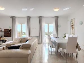 Un salon moderne comprenant un canapé, une table à manger et des chaises, de grandes fenêtres avec des rideaux et une vue sur l’extérieur.