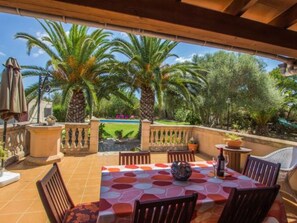 Eine überdachte Terrasse mit Tisch und Stühlen, Palmen und Blick auf Pool und Garten.