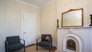 Una habitación con dos sillas, una puerta blanca, una chimenea con un espejo arriba y un tapete estampado en el suelo.