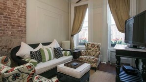 Un salón con pared de ladrillo, un sofá con cojines estampados, un sillón floral, un televisor y una ventana con cortinas.
