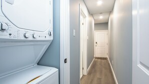 A laundry room with a front-loading washer and dryer, a hallway with wooden flooring, and a door at the end. - Executive 2-Bedroom Suite | Soundproof | Steps to Wortley Village & Vic Hospital (London)