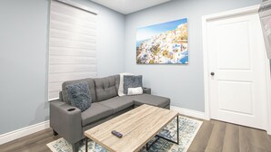 A modern living room with a grey sectional sofa, a wooden coffee table, and a painting of a coastal town. - Executive 2-Bedroom Suite | Soundproof | Steps to Wortley Village & Vic Hospital (London)