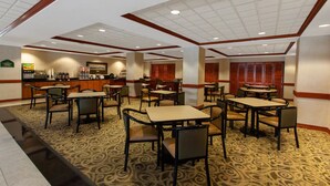 A hotel dining area with multiple tables and chairs, a counter with a beverage station, and patterned carpet flooring. - Hotel Willowbrook (Houston)