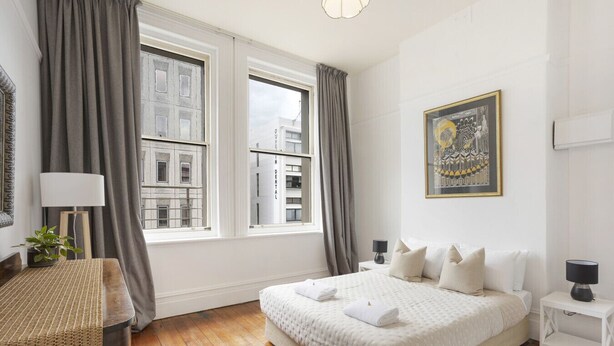 Quarto com uma cama grande, mesa de cabeceira, abajur, quadro na parede e vista para edifícios do lado de fora da janela.