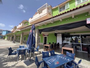 Un espace extérieur aménagé avec des chaises et des tables bleues, un bâtiment vert avec une enseigne, et un ciel dégagé.