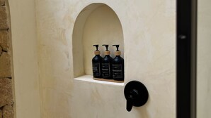 Une salle de bain avec un mur blanc, une petite étagère sur laquelle sont posées trois bouteilles de shampoing et d’après-shampoing, et une poignée de porte noire.