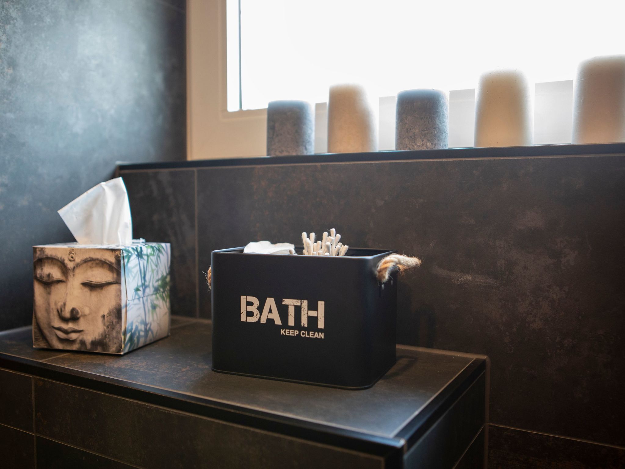 A bathroom counter with a tissue box and a black container labeled "BATH KEEP CLEAN" with cotton swabs.