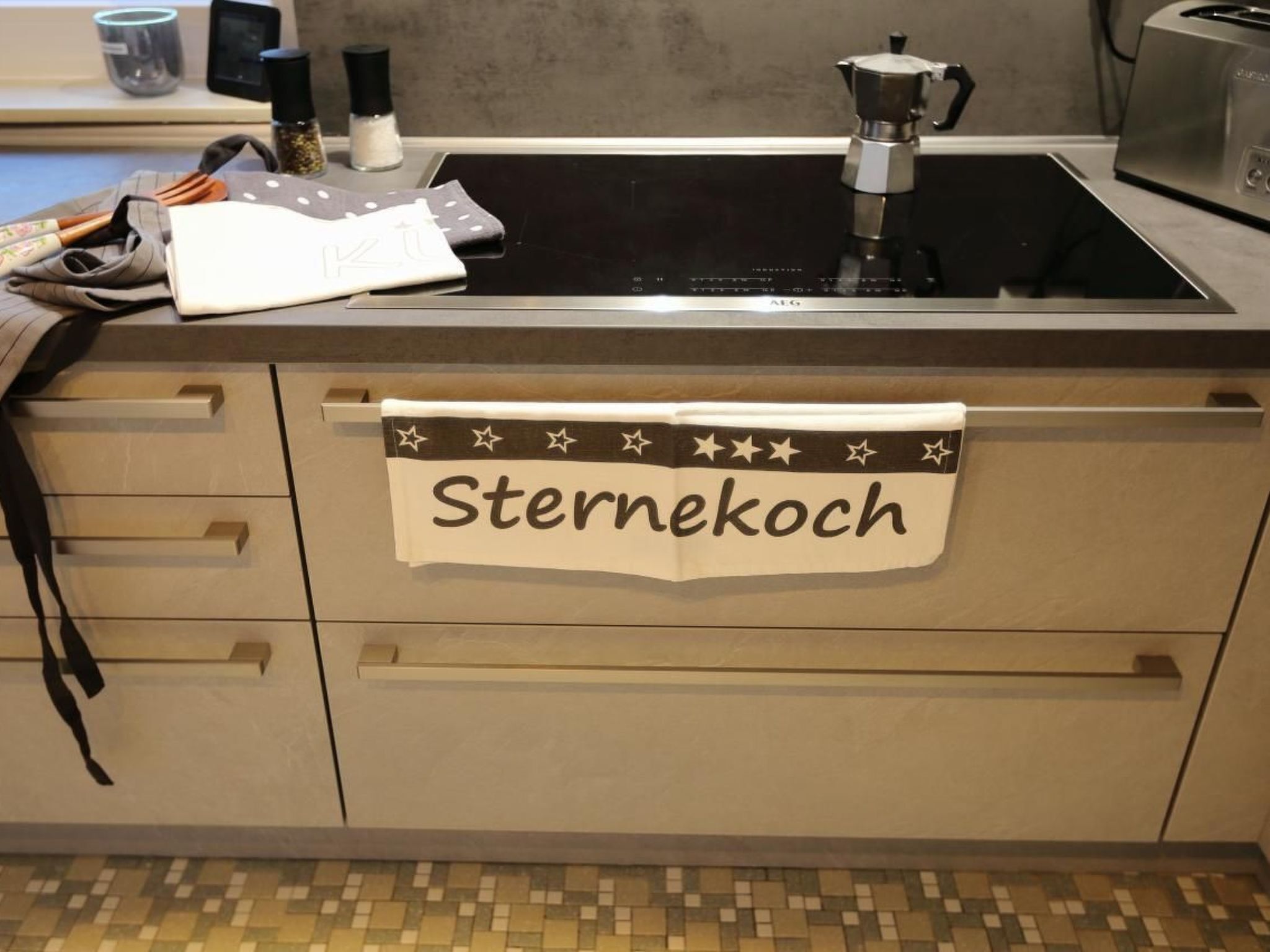 A kitchen with a stove, sink, and a towel hanging on the cabinet that reads "Sternekoch".