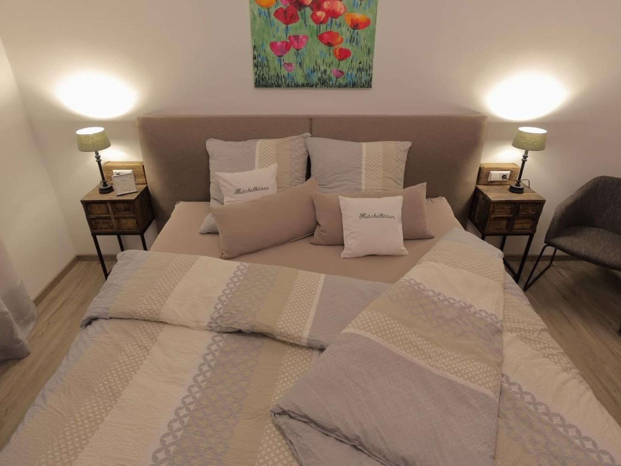 Ein ordentlich bezogenes Bett mit einem gemusterten Überwurf und mehreren Kissen, zwei Nachttische mit Lampen sowie ein Blumenbild an der Wand.