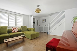 A living room with a green sofa, a wooden coffee table, and a red ottoman. A staircase is visible in the background. - Spacious 3BDR House with Garden in East Ham (Greater London)