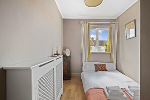 Une chambre à coucher comprenant un lit, une table de chevet, un radiateur, une fenêtre avec des rideaux et une vue sur les bâtiments à l’extérieur.