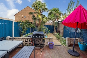 A backyard with a barbecue grill, a wooden deck, a blue fence, and a pink umbrella. - Spacious 3BDR House with Garden in East Ham (Greater London)