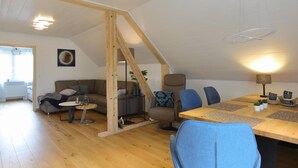 Un amplio loft con techo alto, vigas de madera, un comedor con mesa y sillas de madera, un conjunto de sofás y una escalera.