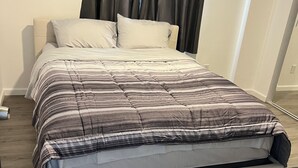 A neatly made bed with a striped comforter and white pillows, a wooden floor, and a black curtain. - Cozy 2-bedroom house in gorgeous Carson with AC for relaxing stays (Carson)