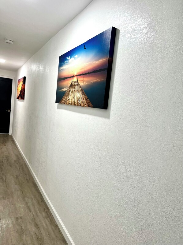 Un couloir aux murs blancs et au plancher en bois. Trois tableaux encadrés sont accrochés au mur, représentant un coucher de soleil sur une jetée, un coucher de soleil sur une montagne et un coucher de soleil sur une étendue d’eau.