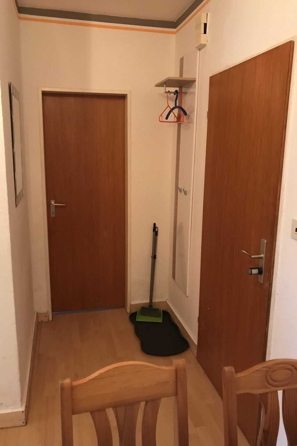 Un couloir avec deux portes en bois, un balai et un porte-manteau avec des vêtements.