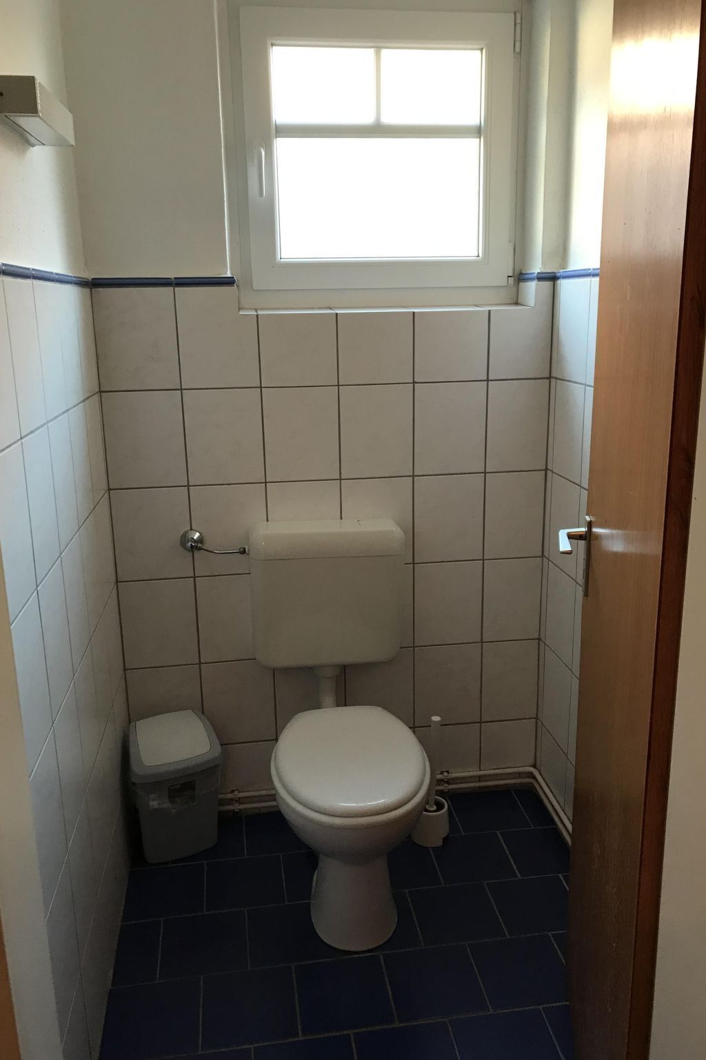 A small, tiled bathroom with a toilet, a small trash bin, and a window above the toilet.
