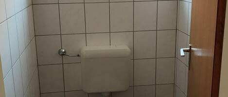 A small, tiled bathroom with a toilet, a small trash bin, and a window above the toilet.