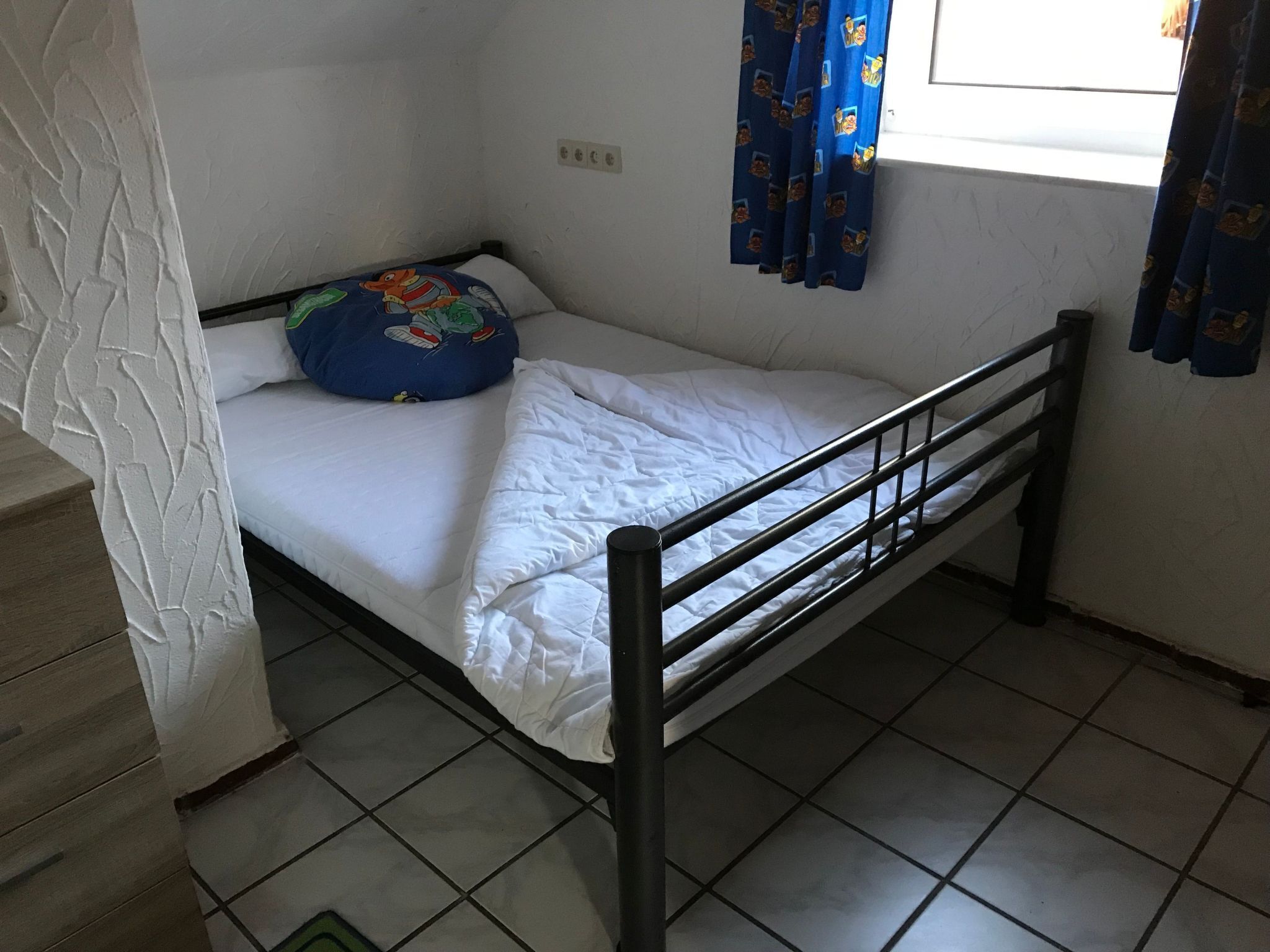 Quarto pequeno com uma cama de solteiro, estrutura de metal, roupa de cama branca, almofada colorida e janela com cortinas azuis.