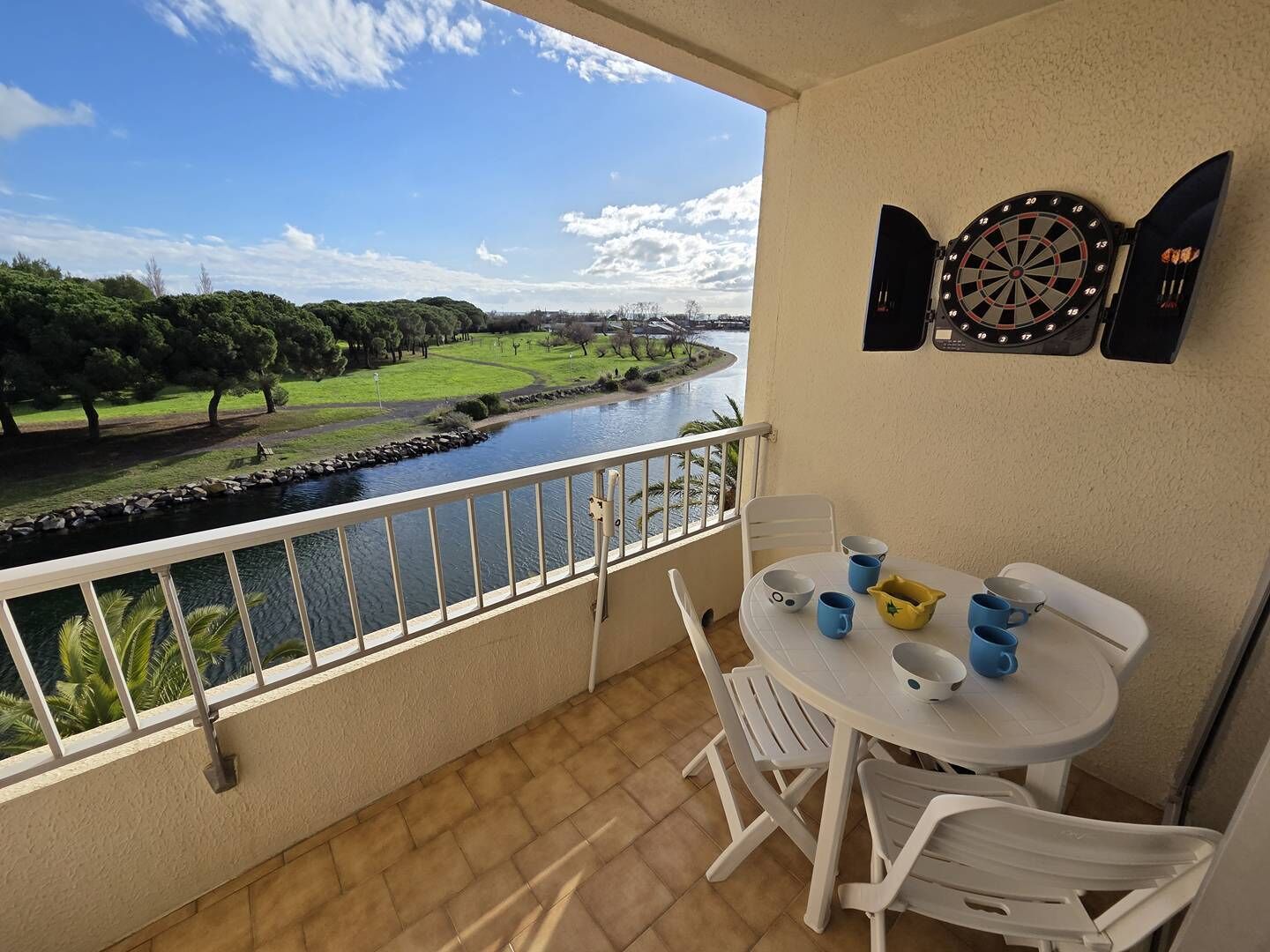A balcony with a view of a river, a dartboard, and a table with chairs and cups.