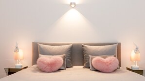 Een net opgemaakt bed met een grijs hoofdeinde, twee hartvormige kussens in roze en twee nachtlampjes, elk met een konijntje erop.