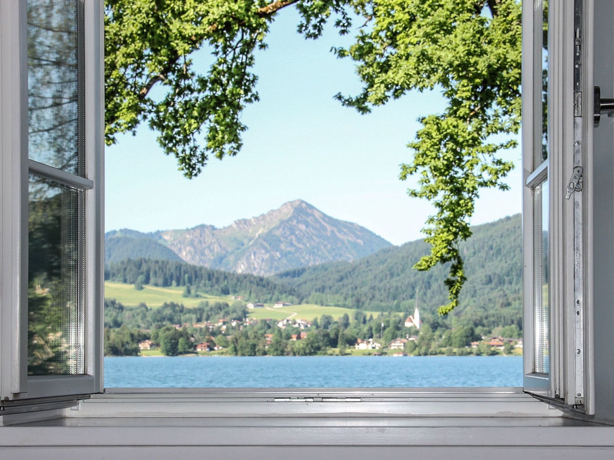 Blick durch ein offenes Fenster auf eine Berglandschaft mit einem See und einem Dorf.