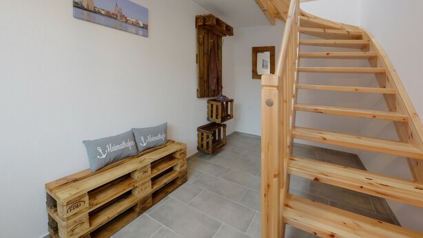 Eine Holztreppe führt zu einer oberen Etage, eine hölzerne Bank mit Kissen und ein gerahmtes Bild an der Wand.