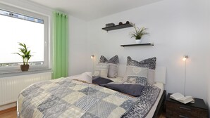 A bedroom with a large bed, a potted plant by the window, a bedside table with a lamp, and a shelf above the bed. - Moderne Wohnungen in historischem Ambiente unweit zur Altstadt und zum Hafen by Interhome (Stralsund)