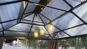 Un patio al aire libre con una mesa de madera y sillas de mimbre, bajo una cubierta de vidrio con luces colgantes.