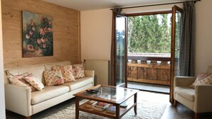 A living room with a sofa, a coffee table with books, a painting on the wall, and a balcony with a view of trees. - Kräuterhof-Ferienwohnung by Interhome (Missen-Wilhams)